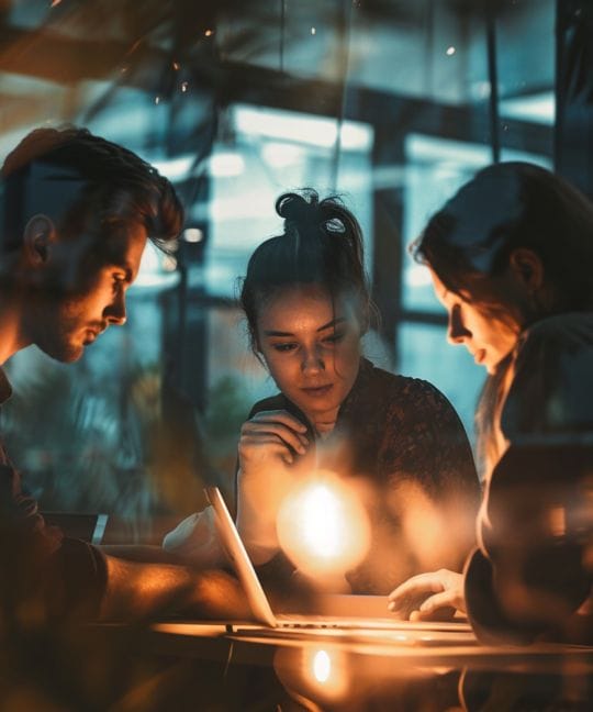 Three people sit around a table engaged in discussion, illuminated by a warm overhead light. They are focused on a laptop screen, with reflections of the room's ambient lights visible on the window behind them.
