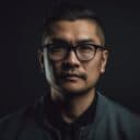 A person with short hair and glasses is looking directly at the camera against a dark background, wearing a dark jacket and shirt. The lighting highlights their face, creating a focused and introspective expression.